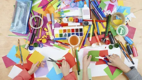 an assortment of paints, markers, paper, glue, scissors and other art supplies shown from above with two sets of hands at the bottom cutting paper.