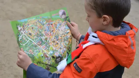 A child in an orange and blue jacket holds a paper map while standing outdoors.