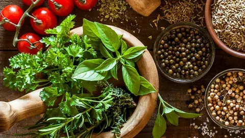 Fresh herbs rest in a mortar beside tomatoes and spices