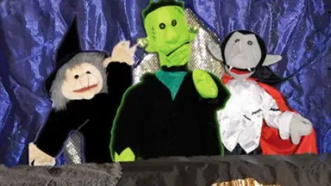 A witch hand puppet, Frankenstein's Monster hand puppet and Dracula hand puppet in front of a purple cloth background.