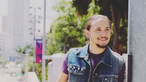 person in denim vest leans against side of building with downtown Los Angeles in background