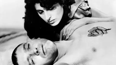 The male protagonist is on bed the female protagonist is bent over him.