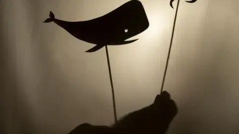 Two hands holding up a shadow puppet of a whale and squid on sticks between a light source and a white fabric to make shadows appear.