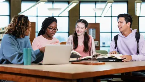 Teens sitting around a table in the library with books and a laptop.