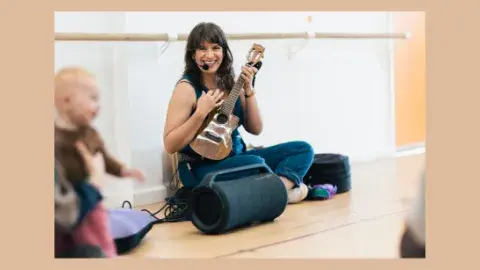 Woman sitting on floor, smiling, and playing ukulele. Baby smiling.