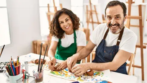 Two adults wearing aprons are smiling and working clay with their hands. The table in front of them has many different types of art supplies.