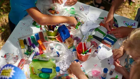 Children crafting with beads, and glue at a table covered with art supplies.