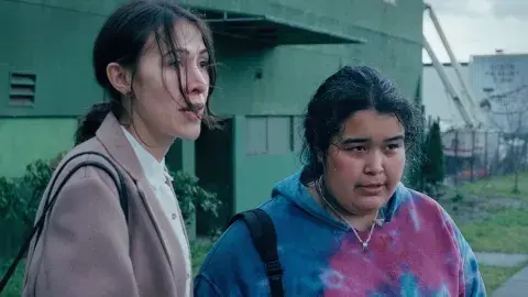 Two indigenous women, one wearing a tie-dye sweatshirt and the other in a more upscale tan coat, stand on a Vancouver street