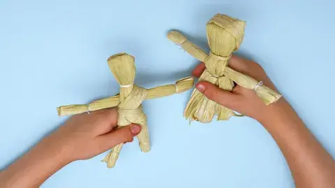 Hands hold two human shaped dolls made out of corn husks