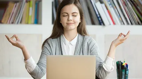 A young woman sitting at a desk with a laptop open. The young woman has her eyes closed and her hands out to her side as if in a meditative state.