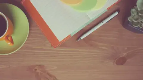 Mug of tea, open notebook, pen, and small plan on top of a wooden surface