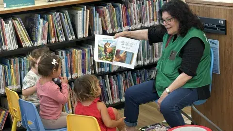 Ms. Judy reads aloud to three children. She is seated in front of a shelf of picture books and is wearing a green STAR volunteer vest.