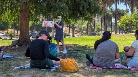masked librarian reads a picturebook to a group of adult caregivers and young children on a lawn under a shady tree