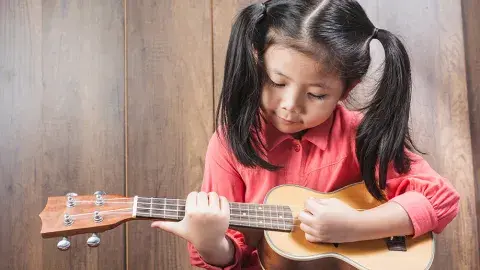 A young girl plays a ukulele in front of a wood-paneled wall.